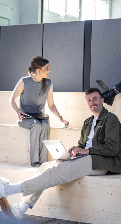 Zwei Kollegen im Gespräch auf einer Holztreppe beim Arbeiten mit einem Apple MacBook.