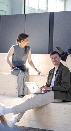 Zwei Kollegen im Gespräch auf einer Holztreppe beim Arbeiten mit einem Apple MacBook.