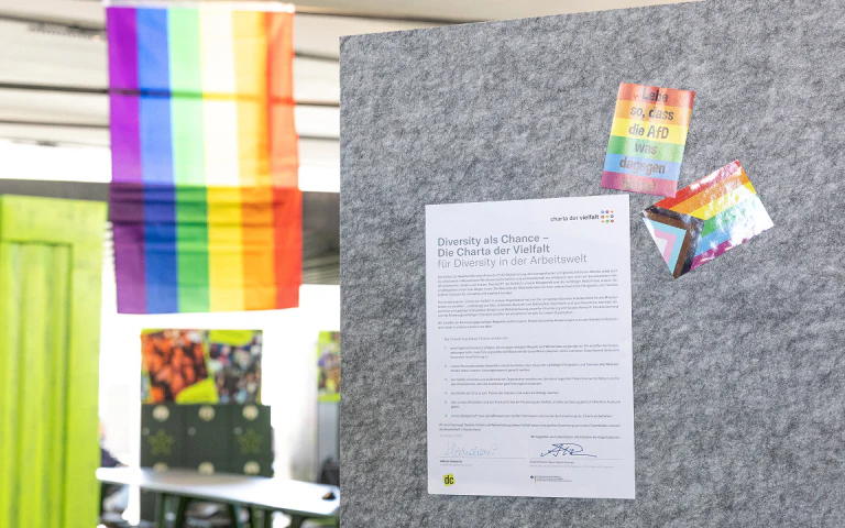 Aushang der Charta der Vielfalt an einer Pinnwand mit Regenbogenflaggen im Hintergrund.