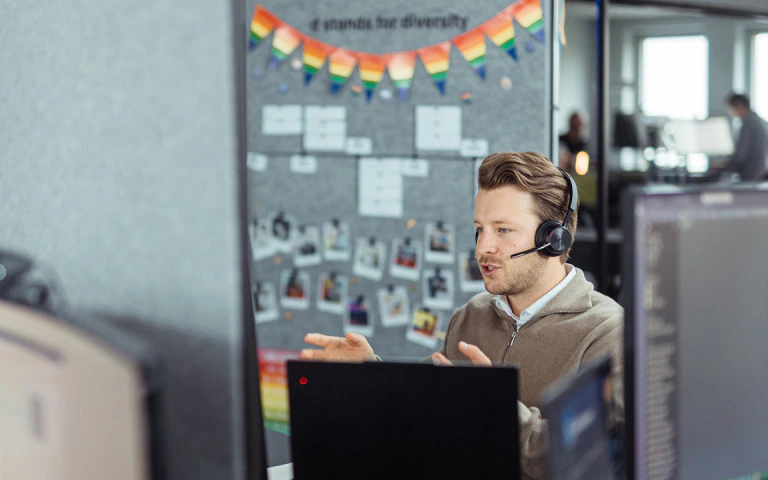 Mitarbeiter mit Headset spricht konzentriert in einem Videocall vor einer Pinnwand mit Regenbogen-Girlande.
