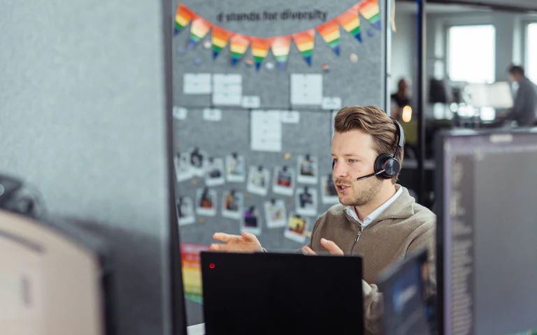 Mitarbeiter mit Headset spricht konzentriert in einem Videocall vor einer Pinnwand mit Regenbogen-Girlande.