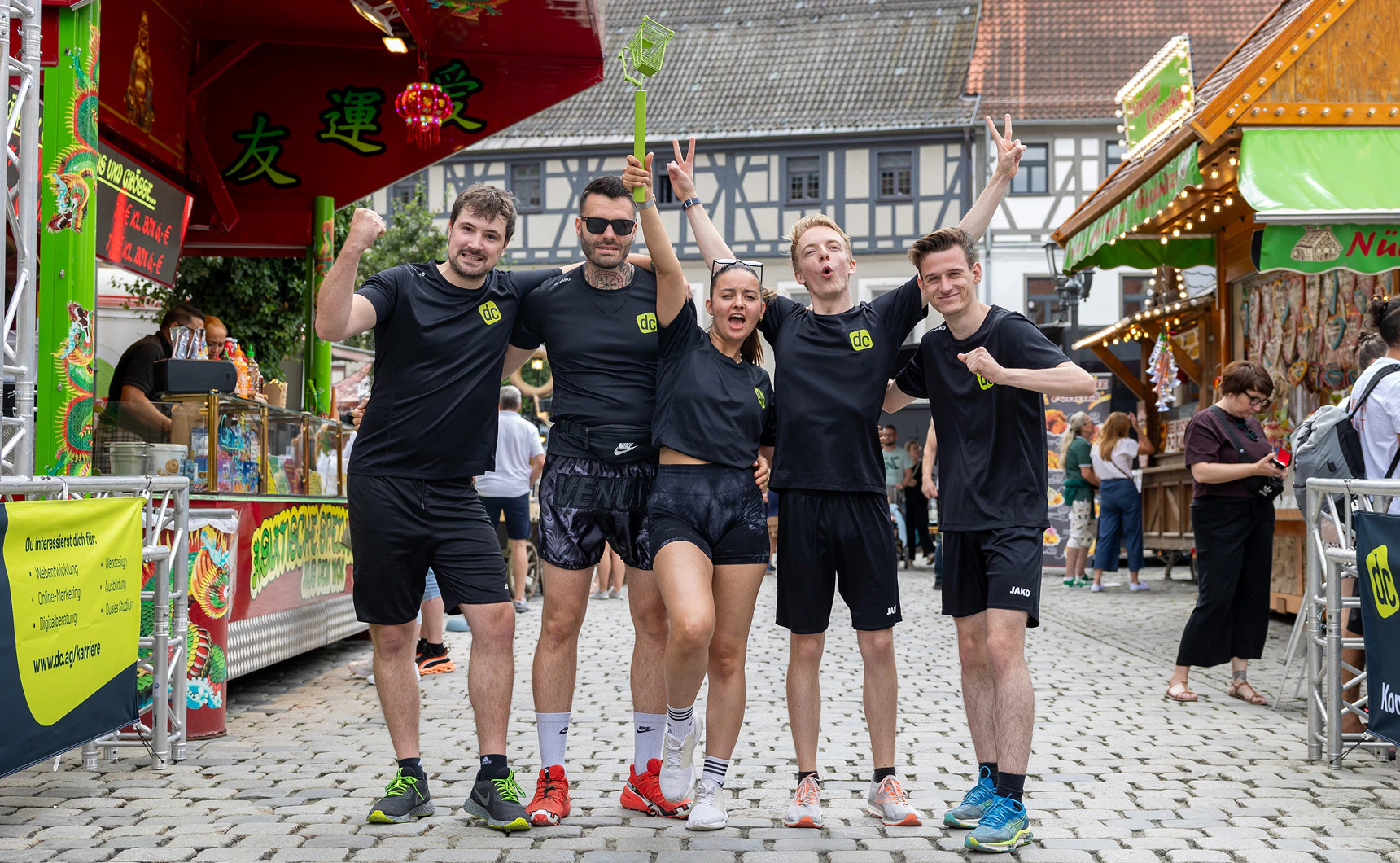 Fünf motivierte Mitarbeiter in schwarzer Sportkleidung von dc posieren jubelnd auf einem Jahrmarkt.