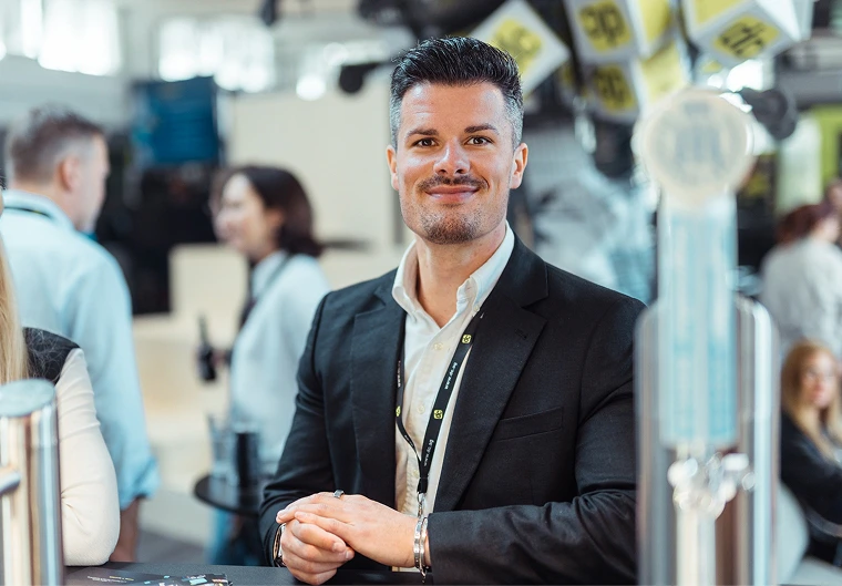 Alexander Streng im schwarzen Blazer lächelt entspannt an einem Messestand, Publikum im Hintergrund