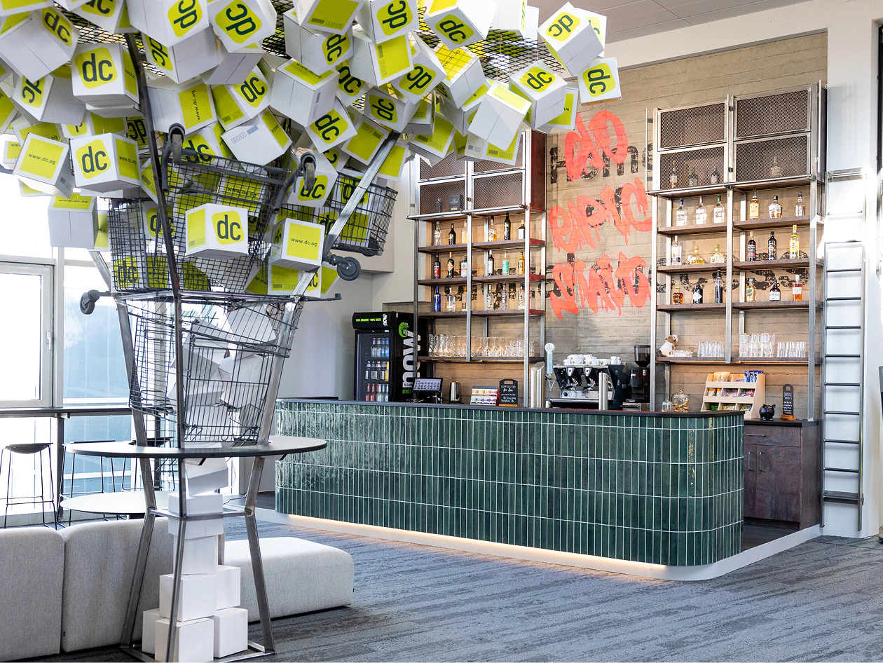 Moderne Cafeteria mit grüner Theke und einer Skulptur aus Einkaufswagen und dc-Kartons im Corporate Design.