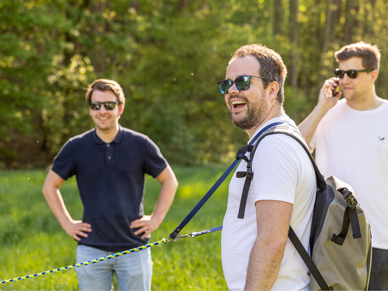 Drei lachende Männer mit Sonnenbrillen stehen auf einer grünen Wiese im Freien.