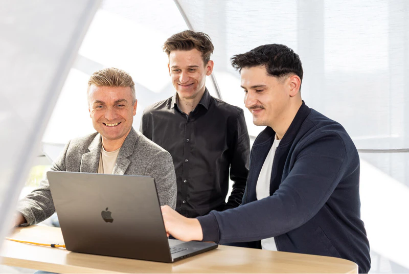 Drei lächelnde Männer besprechen gemeinsam Online-Marketing-Strategien an einem Apple MacBook.