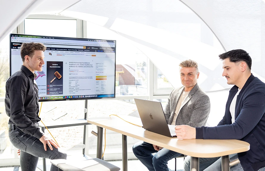 Drei Männer besprechen Amazon-Produktseite für Hundebürste auf großem Bildschirm in modernem Besprechungsraum