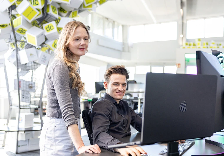 Frau steht lächelnd neben sitzendem Kollegen am Computer im dc-Büro mit gelben Würfel-Dekoelementen