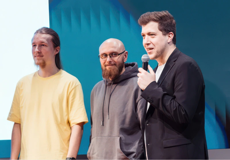 Drei Männer auf Konferenzbühne, einer spricht ins Mikrofon vor blauem Bühnenhintergrund