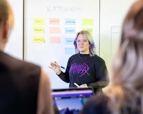 Eine junge Designerin mit schulterlangem, blond-lila Haar, moderner Brille und Metal-Tshirt steht vor einem Whiteboard, das verschiedene Seitenkategorien zeigt.
