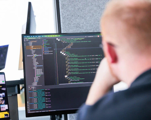 Ein Blick über die Schulter eines rothaarigen Entwicklers mit Brille außerhalb des Fokus zeigt einen Bildschirm, auf dem Code dargestellt wird.
