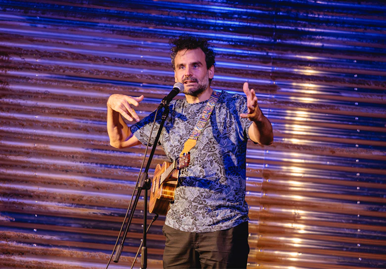 Ein Poetry Slammer mit Ukulele gestikuliert während eines Auftritts am Mikrofon.