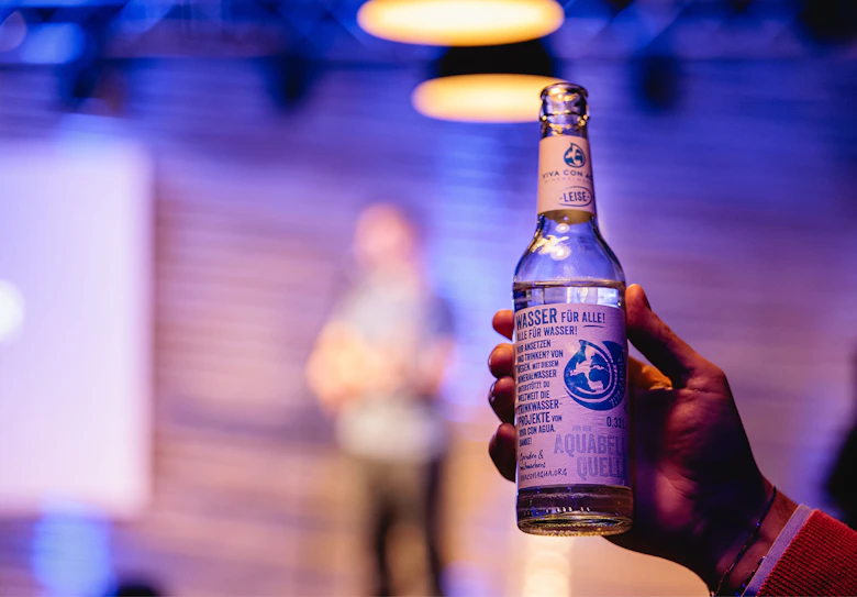 Hand hält eine Flasche Viva con Agua Wasser vor einer Poetry-Slam-Bühne.