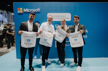 Vier lächelnde dc AG Mitarbeiter präsentieren weiße dc AG Jutebeutel vor einer blauen Microsoft-Wand auf der Directions EMEA 2024.