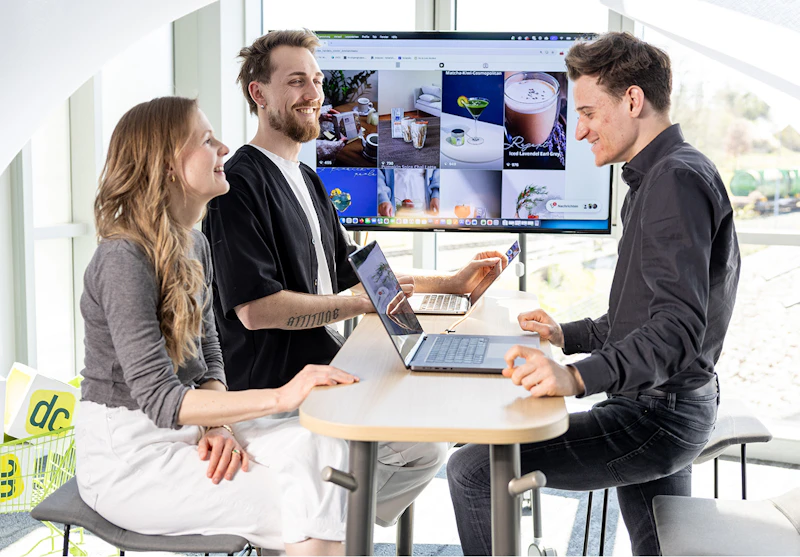 Drei Personen besprechen Content-Strategie mit Laptops vor großem Bildschirm mit Produktfotos im dc-Büro