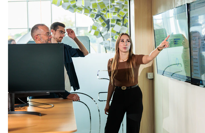 Frau zeigt zwei Kollegen bei einer Digitalberatung Daten auf einem großen Monitor.