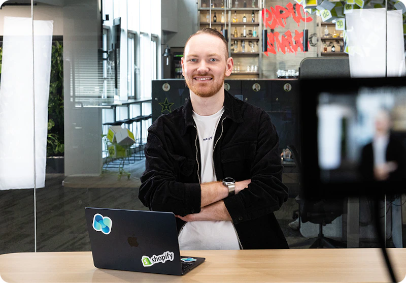 Lächelndes dc AG Teammitglied steht mit verschränkten Armen hinter einem Apple MacBook mit Shopify-Sticker.