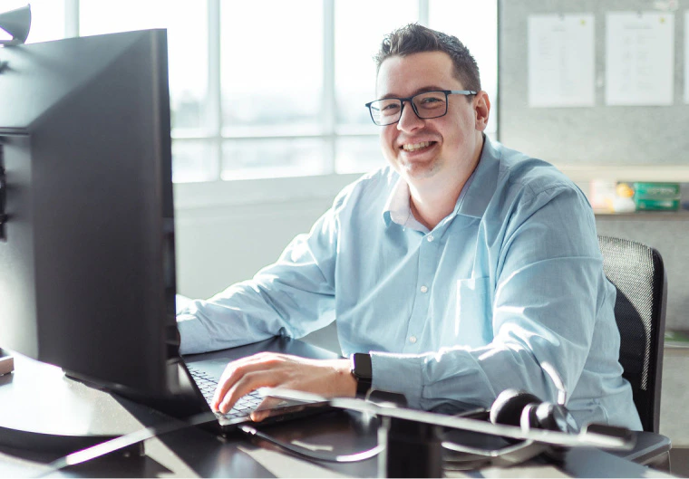 Lächelnder Marcel Schmid arbeitet am PC-Monitor und Laptop im hellen Büro.