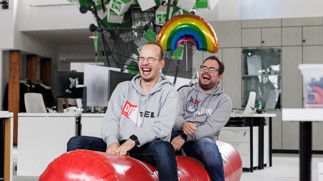 Zwei lachende Mitarbeiter der dc AG sitzen auf einem roten Boxsack unter einem Regenbogen-Luftballon.
