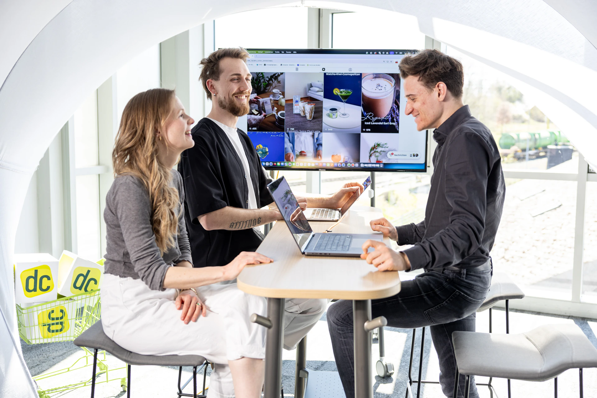 Drei Menschen besprechen eine Online-Marketing-Strategie an Laptops vor einem großen Monitor.