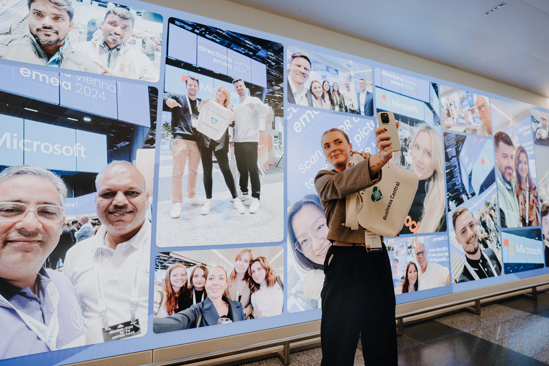Frau macht ein Selfie vor einer Fotowand der Directions EMEA 2024 mit Microsoft-Logo.